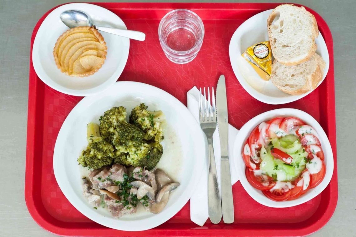 French school lunch tray with chicken and broccoli, tomato and cucumber salad, fresh bread and camembert cheese and apple tart served in a French school cafeteria lunch