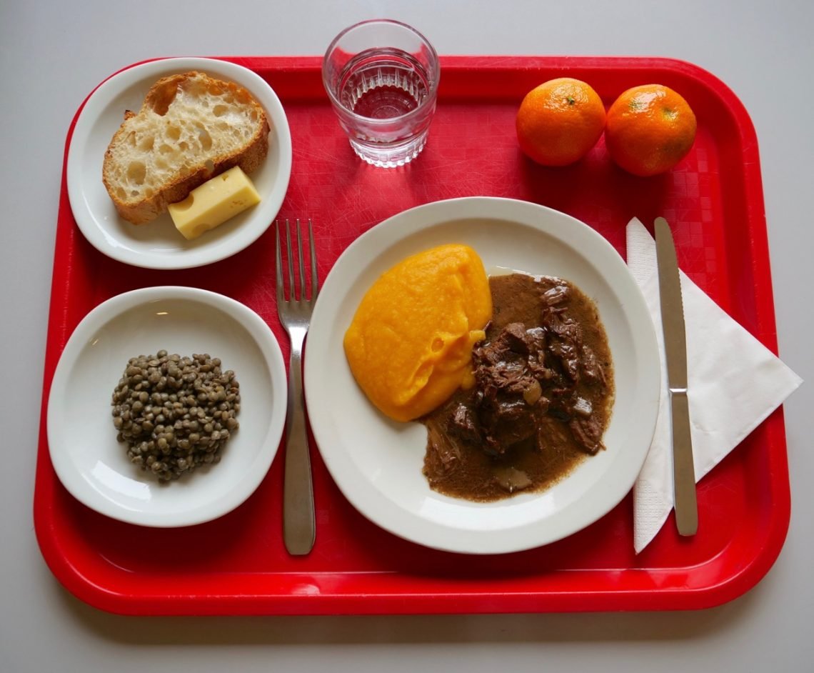 French school lunch tray with beef stew, carrot purée, green lentils, bread, cheese, water, and clementines in a public elementary school cafeteria.
